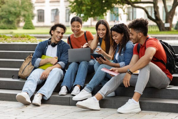 Diverse group of smiling college students sitting on steps outside and looking at a laptop