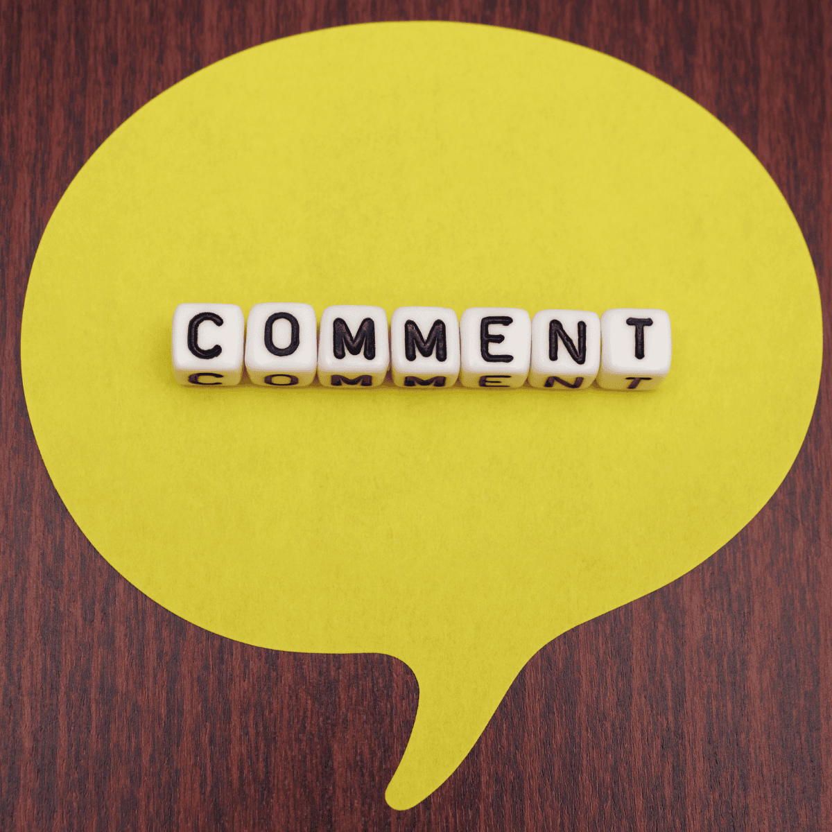 Yellow speech bubble shape on a dark wood background with the word “COMMENT” spelled in white and black letter tiles across the center.