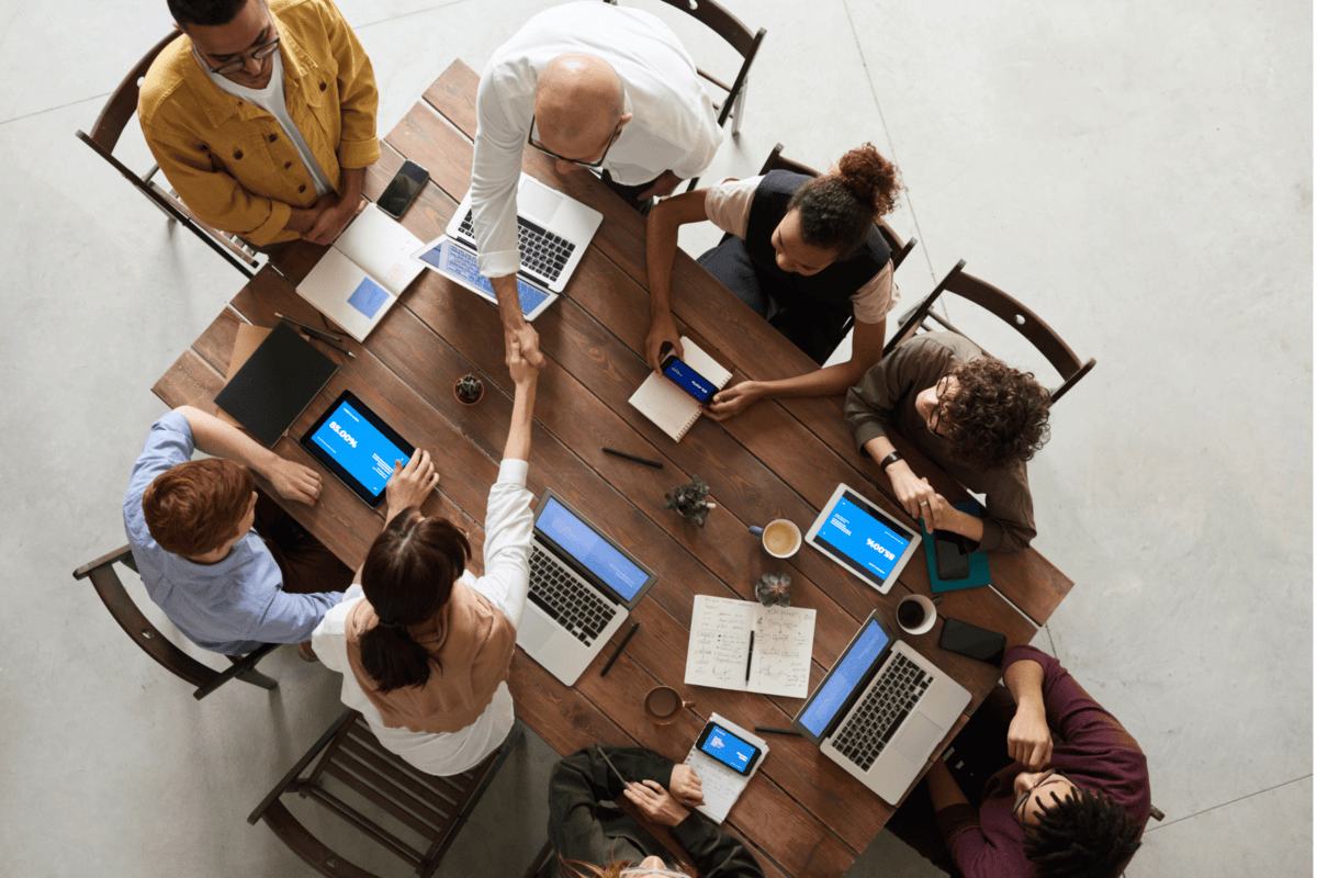 View from above of a group of meeting attendees around a table, with laptops, pads and pencils.