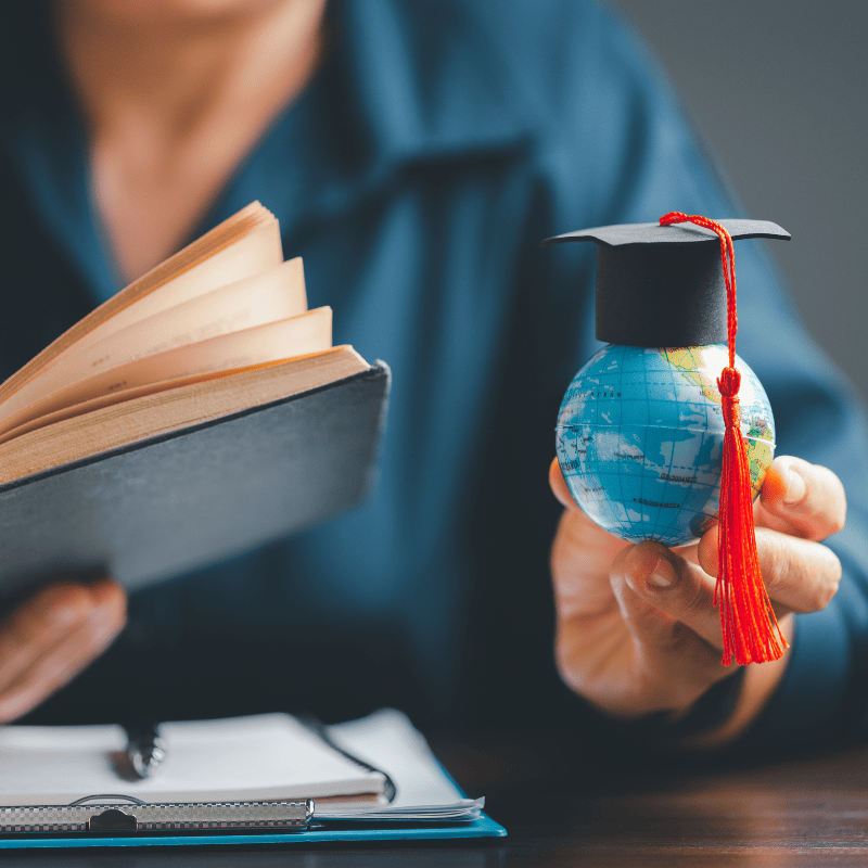 A person at a desk holding a book and a globe with a graduation cap on it