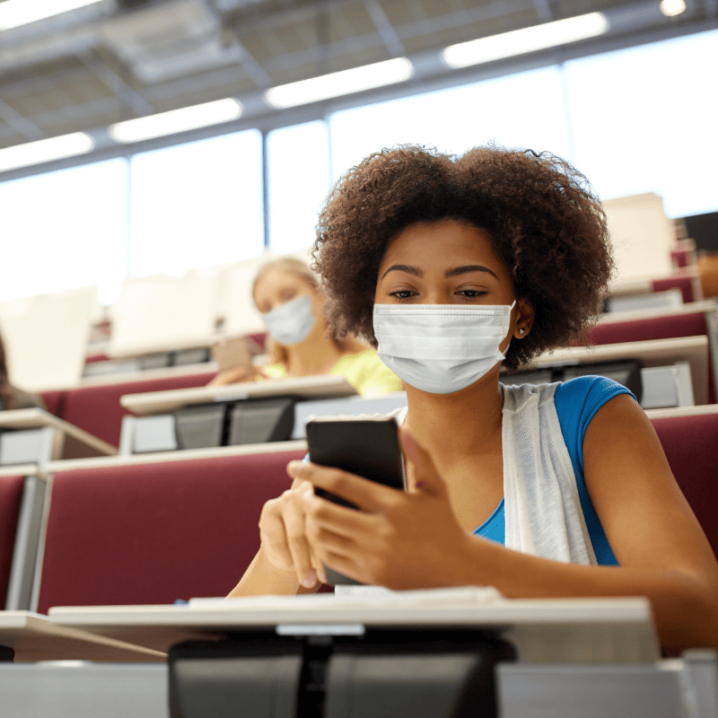 A college student wearing a face mask while using her phone in a lecture hall