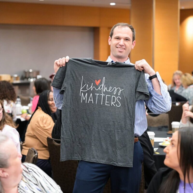 A smiling conference attendee holding up a T-shirt that says Kindness Matters