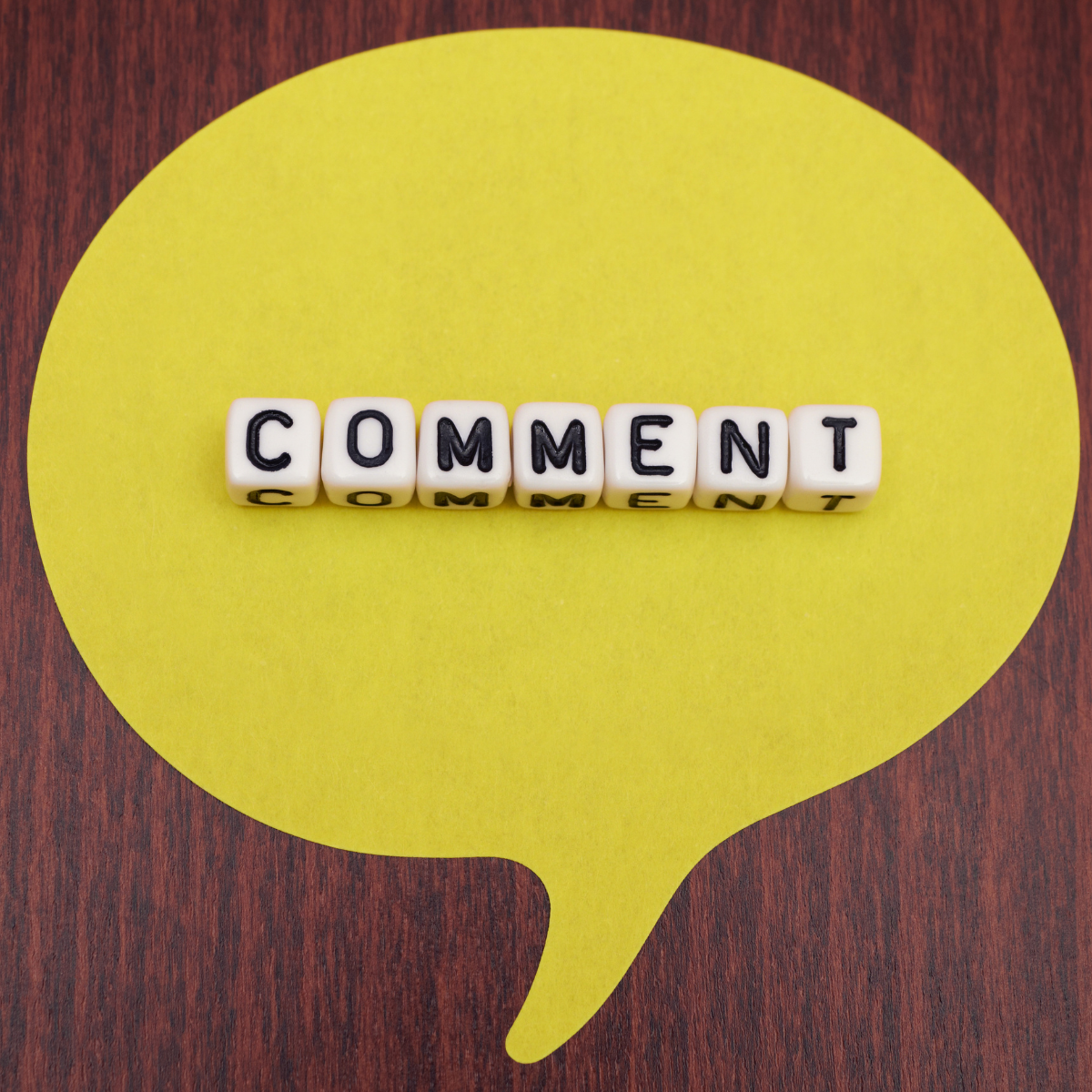 Yellow speech bubble shape on a dark wood background with the word “COMMENT” spelled in white and black letter tiles across the center.