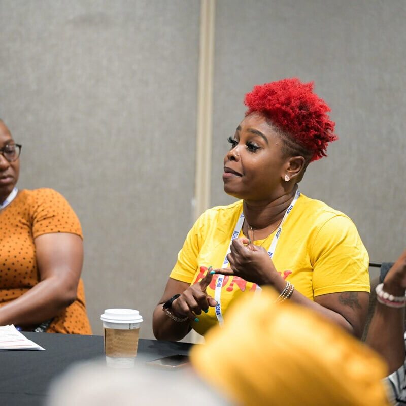 A conference attendee with bright red hair speaking to other attendees at a table.