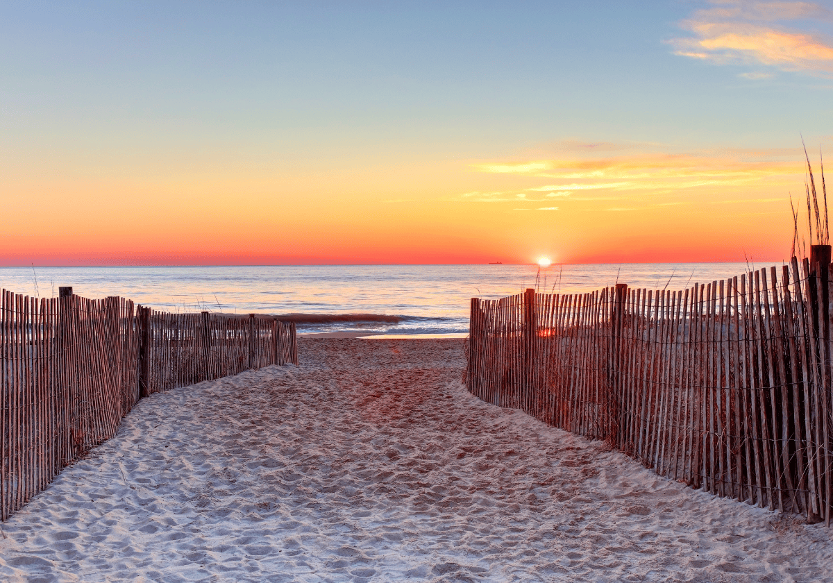 Sunset at Rehoboth Beach, Delaware - a fenced path in the sand leading out to the shore, with the sun rising over the horizon.