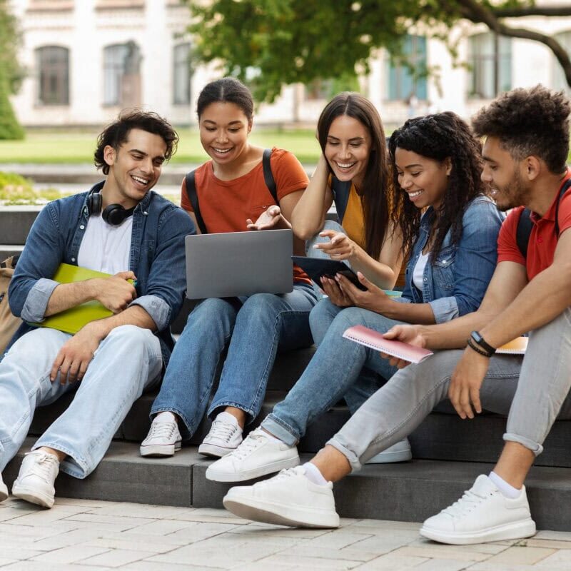 Diverse group of smiling college students sitting on steps outside and looking at a laptop