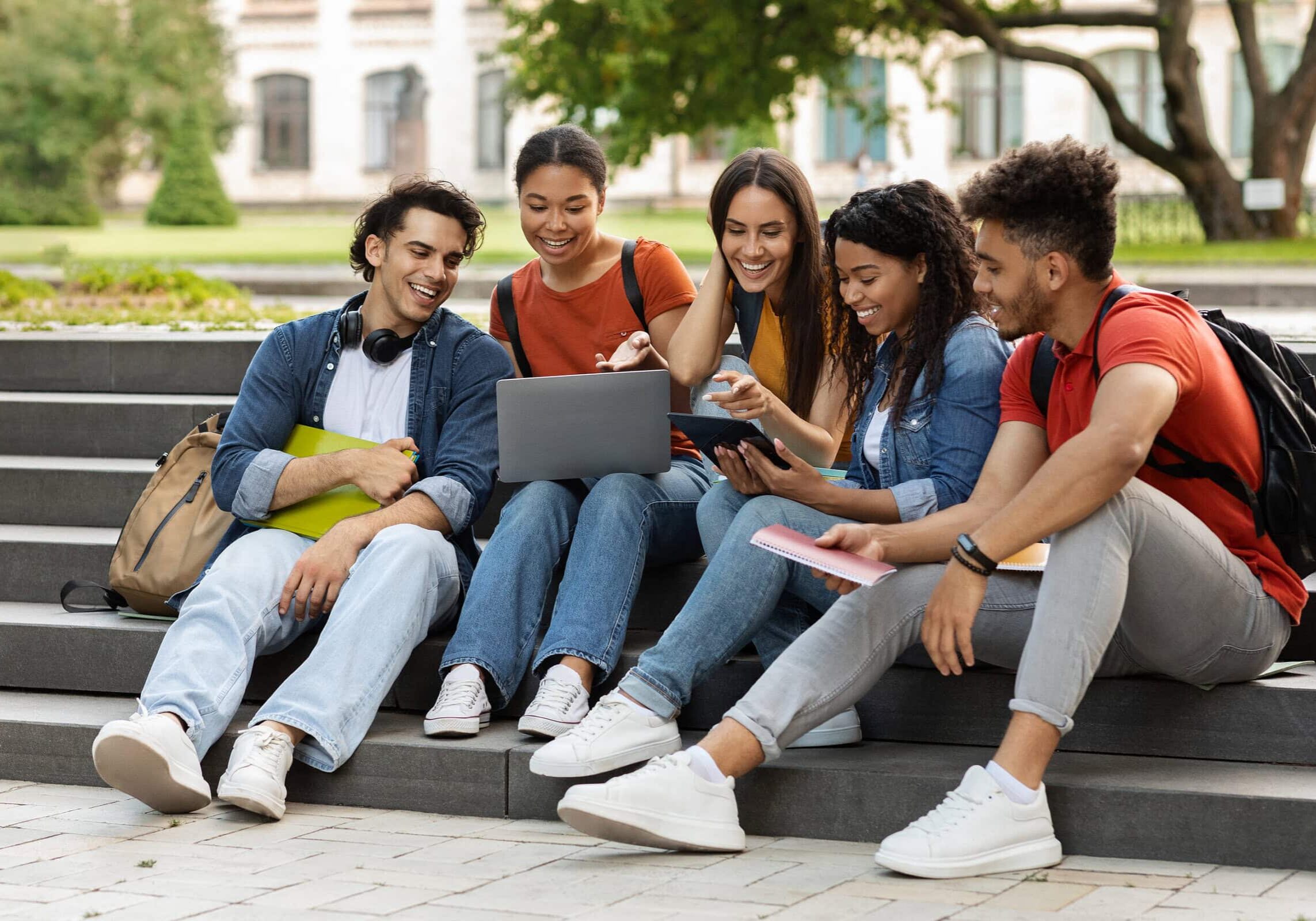 Diverse group of smiling college students sitting on steps outside and looking at a laptop