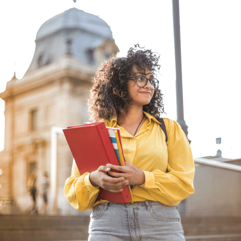 Smiling curly-haired female college student wearing glasses and standing outside holding school books
