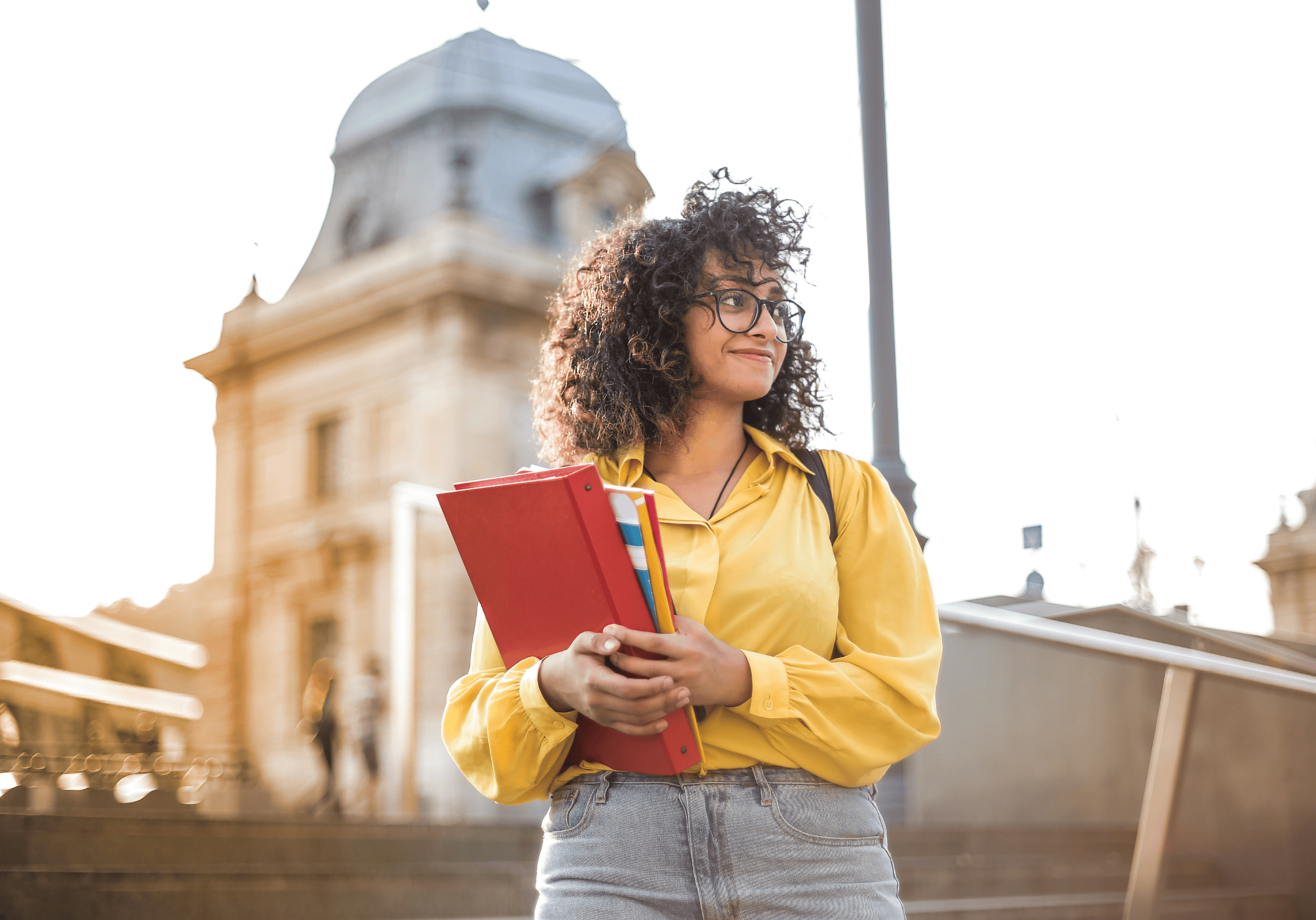 Smiling curly-haired female college student wearing glasses and standing outside holding school books