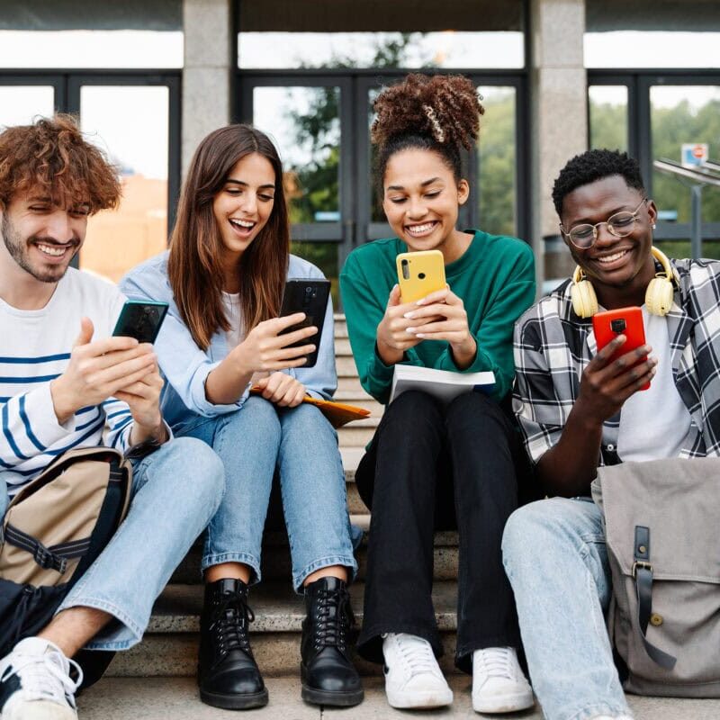 Smiling university student friends using mobile devices together,