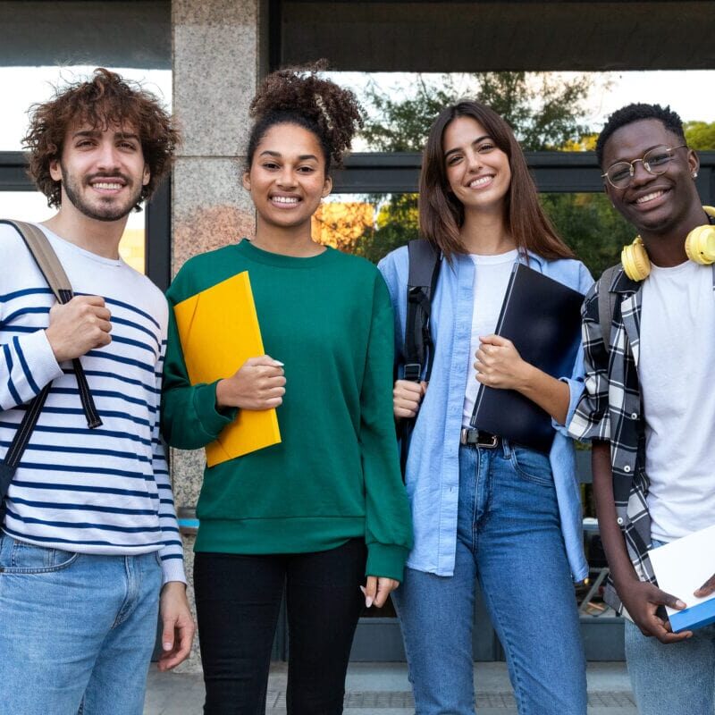 A diverse group of smiling college students standing in front of a building and holding school books