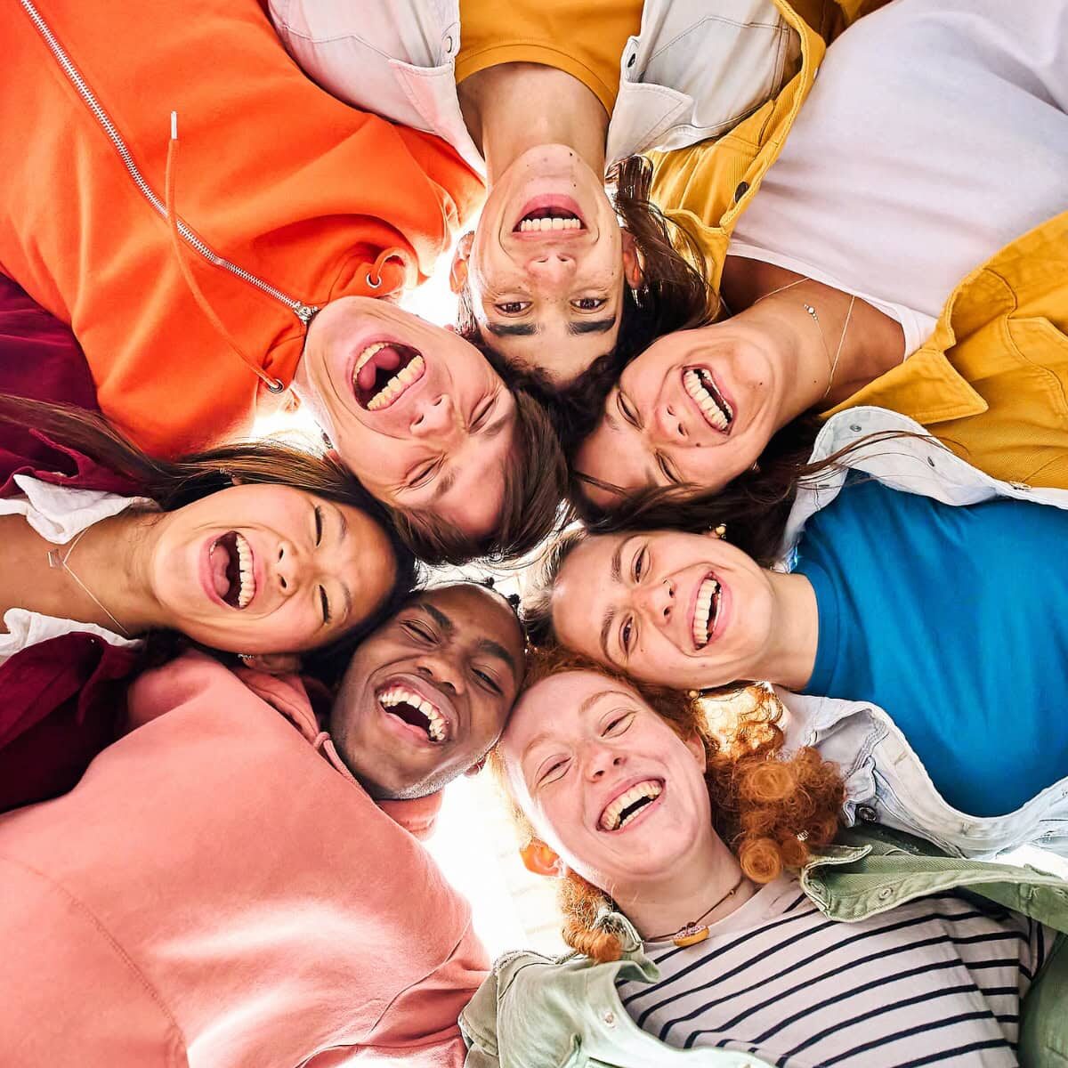 A group of smiling young adults standing in a circle and putting their heads together to look down at the camera
