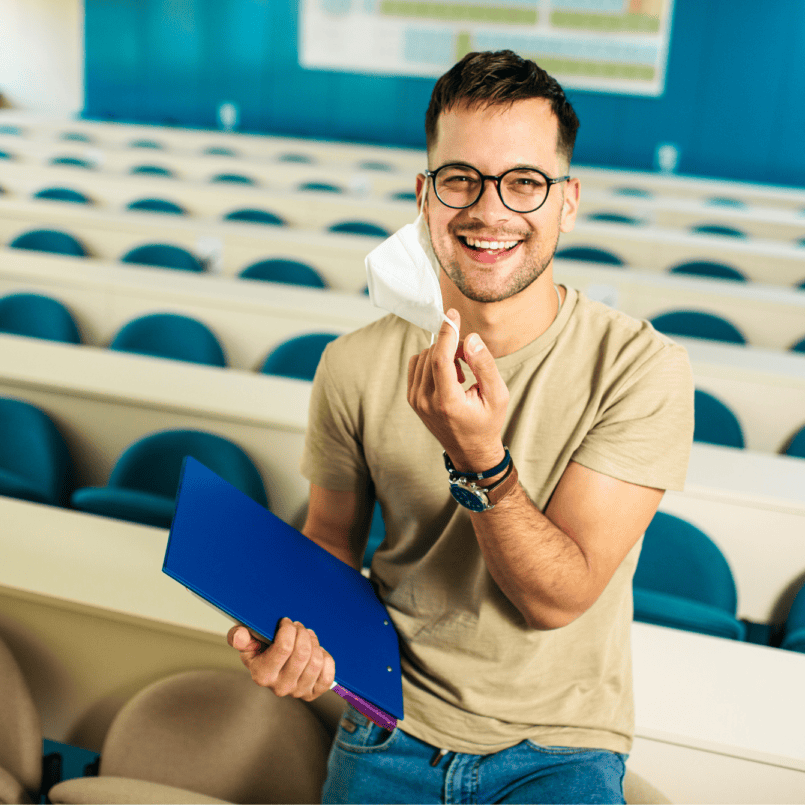Smiling male college student wearing glasses and a face mask and standing in a lecture hall