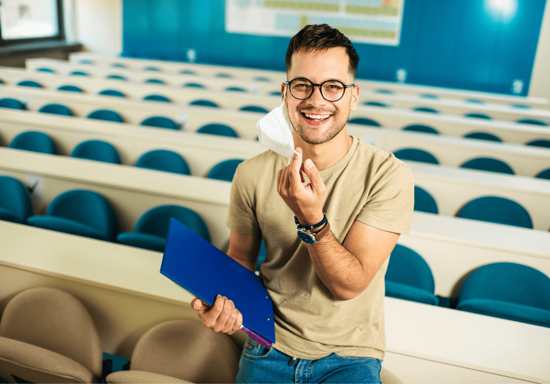 Smiling male college student wearing glasses and a face mask and standing in a lecture hall