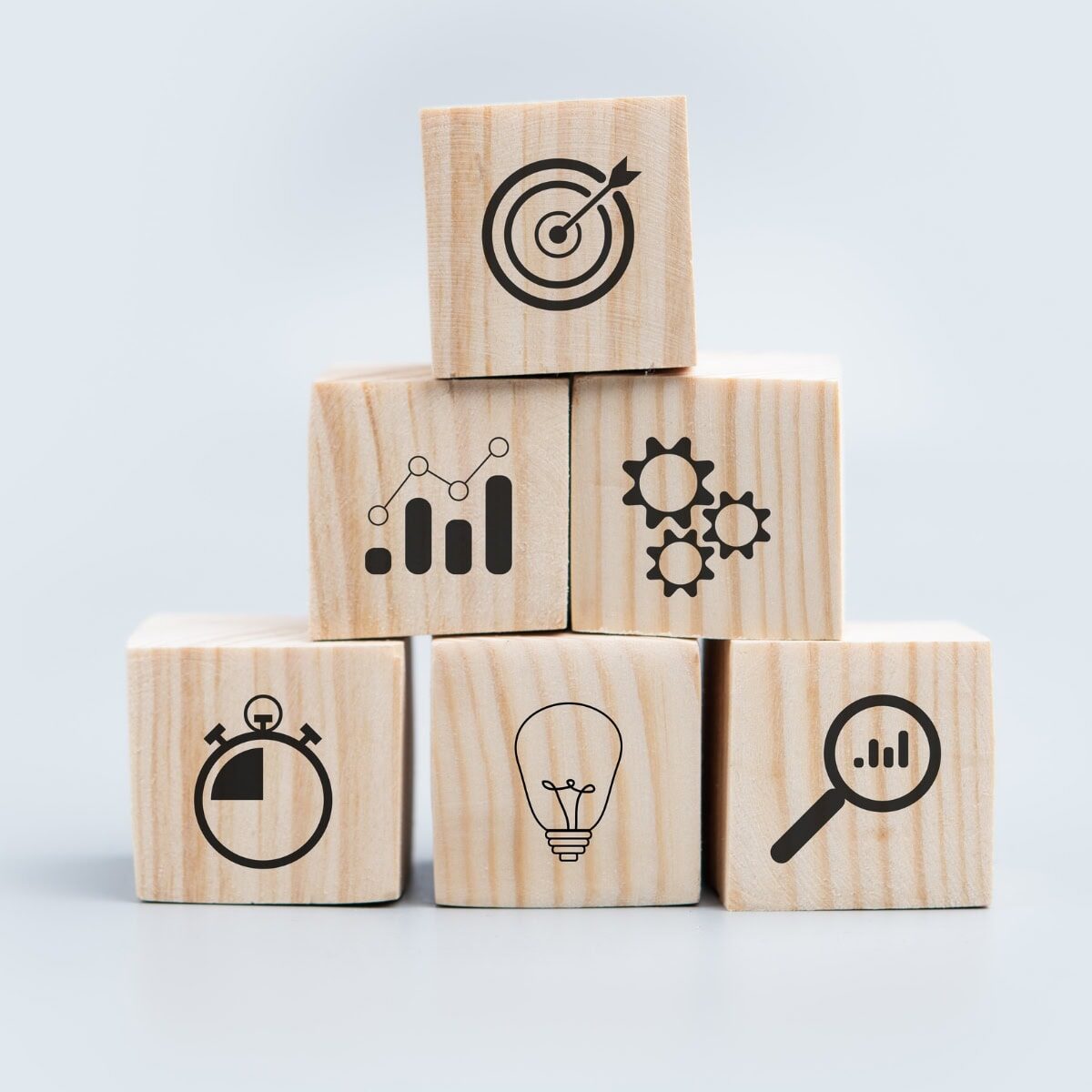 Stacked wooden blocks with various icons: a target, data graph, gears, a stopwatch, a lightbulb, and a magnifying glass
