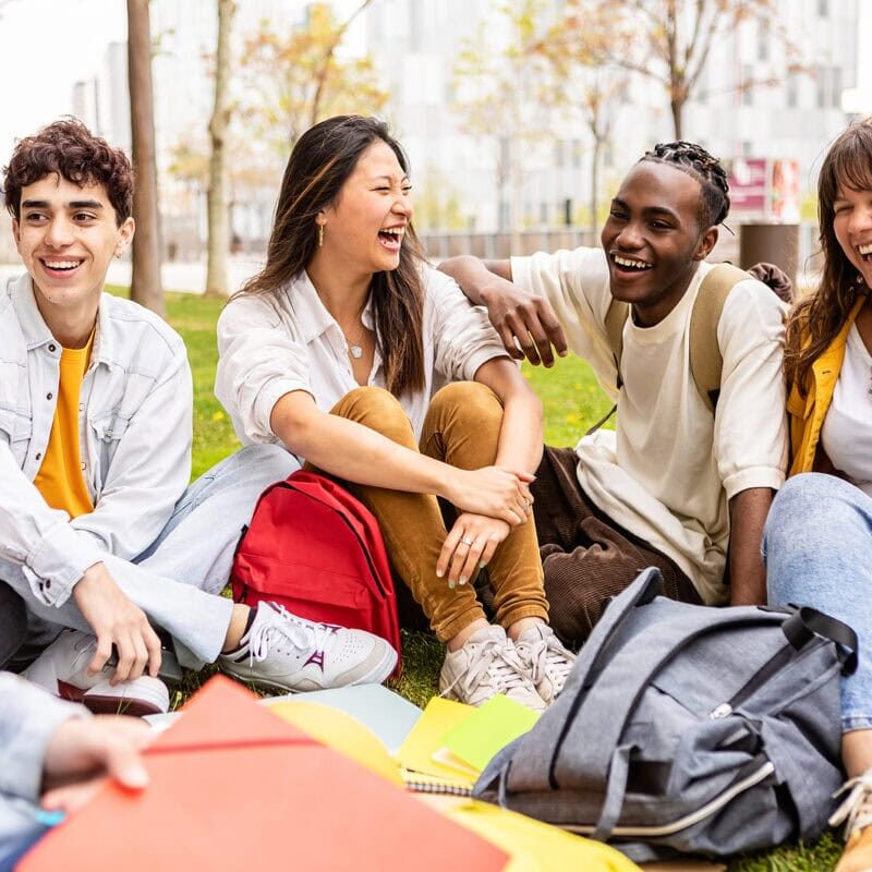Young group of multiracial students laughing and having fun together sitting on the grass in the university campus. Teenage classmates social gathering relaxing outdoors.