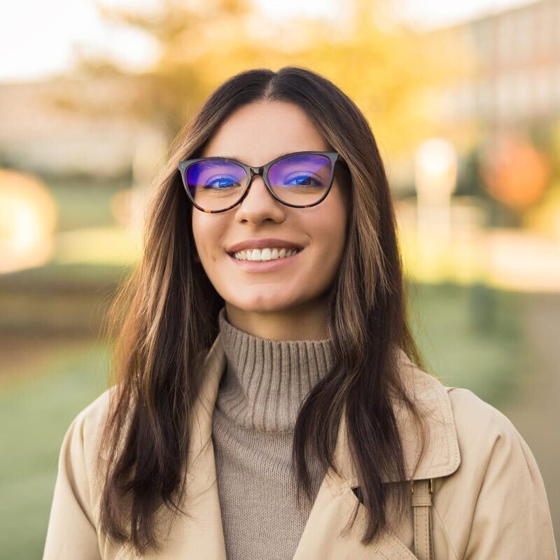 Young woman with glasses and wearing a turtleneck sweater and jacket standing outside in front of fall foliage.