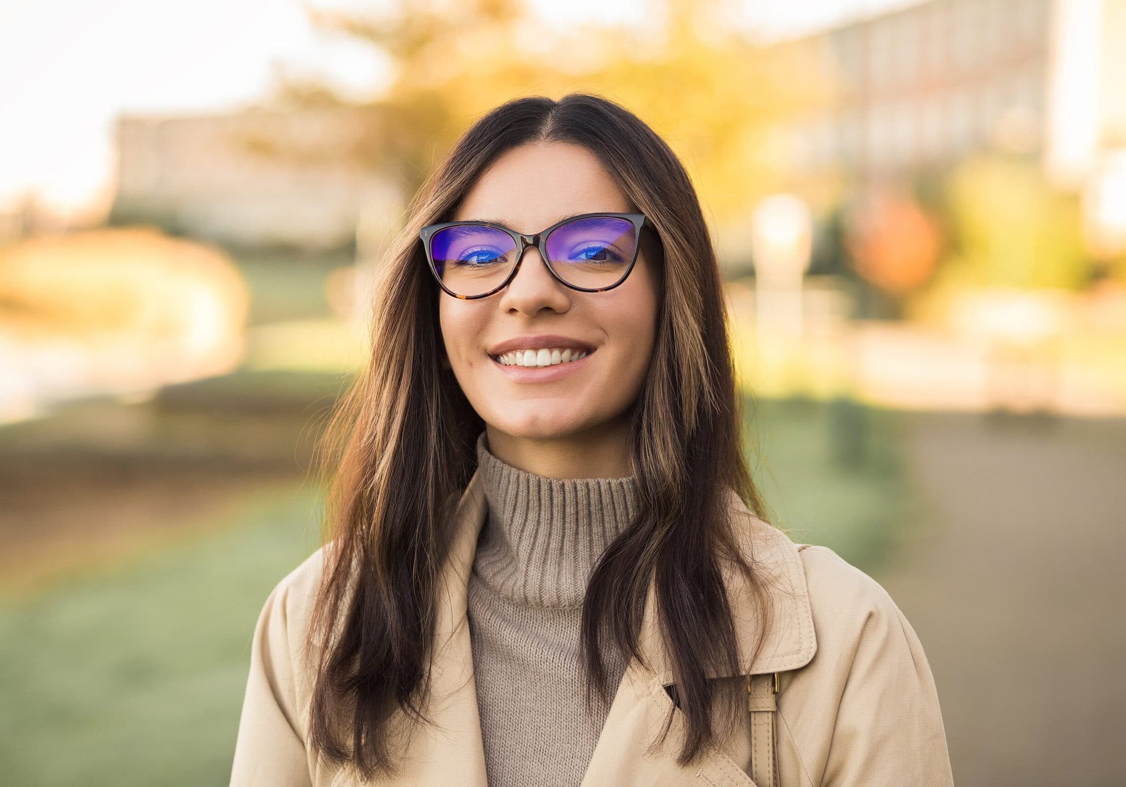 Young woman with glasses and wearing a turtleneck sweater and jacket standing outside in front of fall foliage.