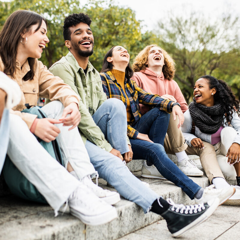 Multiracial group of young teenage student friends laughing together and having fun sitting on city street - United diverse millennial people celebrating together outside - Unity and community concept